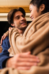 Wall Mural - Smiling man hugging asian girlfriend in blurred blanket outdoors