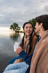 Wall Mural - smiling asian woman holding cup near boyfriend and lake
