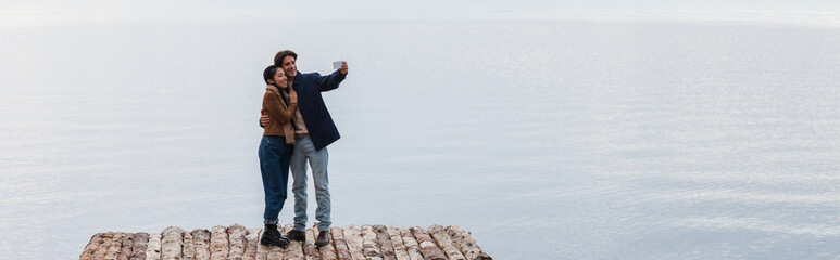 Wall Mural - cheerful interracial couple taking selfie on pier near sea, banner