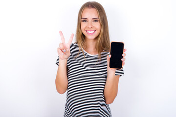 beautiful blonde girl wearing striped t-shirt on white background holding modern device showing v-sign