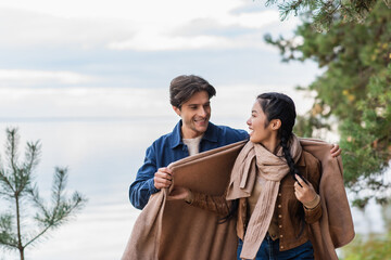 Wall Mural - man holding blanket near asian girlfriend with lake on background
