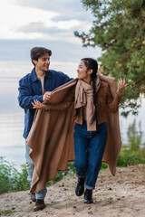 Wall Mural - smiling man holding blanket near asian girlfriend walking on beach