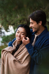 Wall Mural - Smiling man touching cheek of asian girlfriend in blanket outdoors