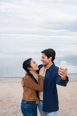 Wall Mural - smiling interracial couple hugging and taking selfie on beach