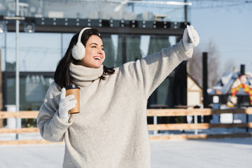 happy woman in sweater and ear muffs holding paper cup and taking selfie on ice rink