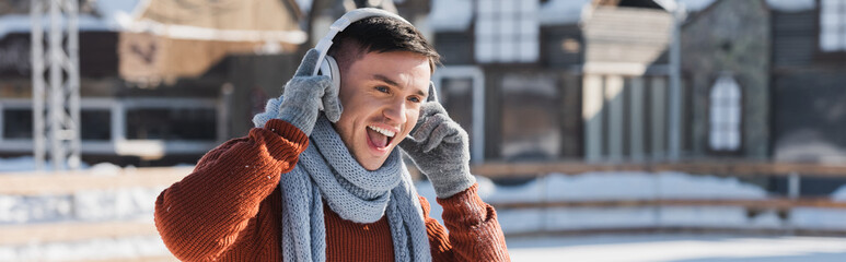 positive young man in sweater and scarf listening music while singing outside, banner