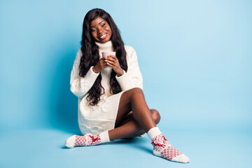 Wall Mural - Photo of dreamy cute dark skin woman dressed white winter clothes sitting floor drinking tea isolated blue color background