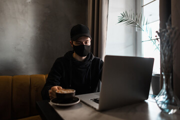 Wall Mural - Young guy with a medical protective mask and a black cap in a hoodie drinks coffee and looks at a laptop in a cafe