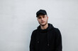 © alones - Fashionable handsome young hipster man in stylish black mock up cap with hoodie stands near gray wall on the street