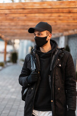 Wall Mural - Stylish handsome man with a protective mask in fashionable clothes with a jacket, hoodie, mockup cap and backpack walks in the city. Men's urban clothing style