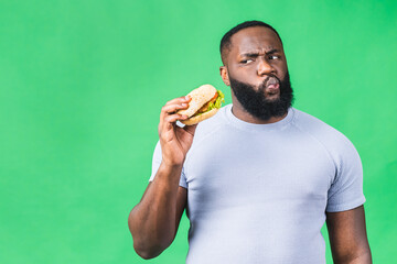 Hungry young african american black man eating hamburger isolated over green background. Diet concept.