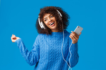 Wall Mural - Joyful and carefree lovely african-american female model in winter sweater, dancing to rhythm music, listening new track on song app, christmas playlist, wear headphones hold smartphone