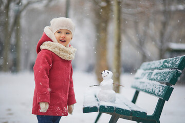 Adorable toddler girl building a snowman on a day with heavy snowfall