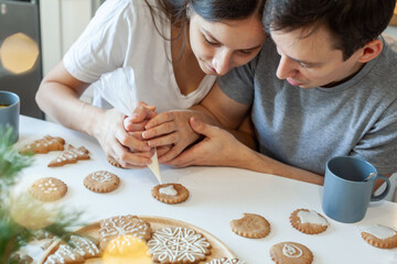 Beautiful man and woman decorate Christmas gingerbread. Two people draw a heart on the cookie. Stylish home kitchen