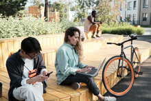 Group Of Three Young Multi-ethnic People Spending Summer Day Outdoors Sitting In Modern Urban Park Working Remotely Or Surfing Internet On Their Gadgets