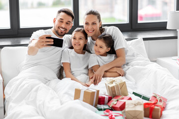 Poster - family, winter holidays and people concept - happy mother, father and two daughters with christmas gifts taking selfie on smartphone in bed at home