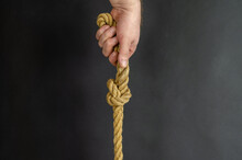 Adult Male Holds Vertically A Rope With A Knot. Hand Holds A Yellow Jute Rope Against A Black Background. Close-up. Selective Focus.