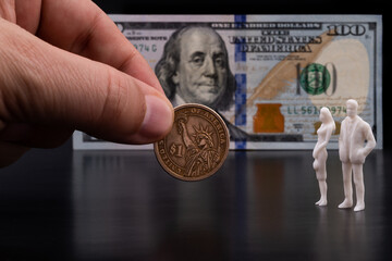 Male fingers are holding out a 1 American dollar coin to two plastic figures of symbolic people against the background of a 100 American dollar bill