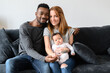 Portrait of happy mixed-race family of three African-American husband, caucasian wife and a cute biracial daughter sitting on the couch indoors and looking at the camera