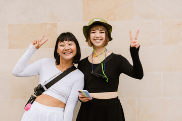 Multiracial two women using cellphone and showing peace gesture