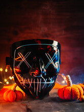 Scary Halloween Neon Blue Mask On A Dark Smoky Background, Knitted Orange Pumpkins, Bokeh Garlands.