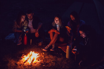 A group of friends are sitting around a campfire at night using smartphones.