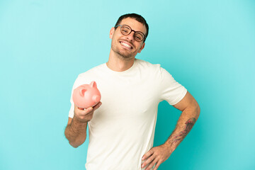 Brazilian man holding a piggybank over isolated blue background posing with arms at hip and smiling