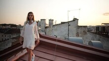 Attractive Blonde Girl In A White Dress Relaxing On A Roof At Sunset, Wind Shuffles Her Beautiful Long Hair. Portrait Of Gorgeous Woman On Rooftop. Urban Cityscape View