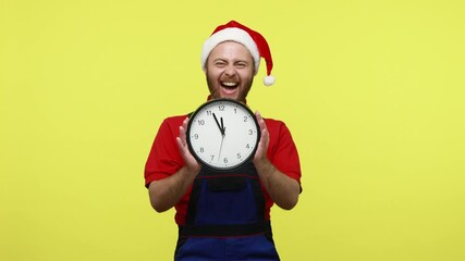 Wall Mural - Happy positive man showing clock and being glad to finish work till New Year Eve, wearing blue overalls, red T-shirt and santa claus hat. Indoor studio shot isolated over yellow background.