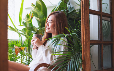 A young woman drinking hot coffee and relaxing on balcony with houseplants garden at home