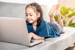 © tai - An adorable smiling child girl on a sofa while online studies on a laptop at home due to the Covid-19 coronavirus pandemic. Online education learning gaming technology and homeschooling concept idea