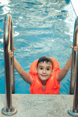 Happy delightful kid with wet hair standing on chrome handrail in fresh water swimming orange outdoors pool lifejacket in summer holiday leisure day. Summer activities for happy child on the pool