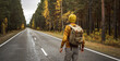 Traveler with a backpack is walking on an empty road along forest. Concept of freedom, travel, hiking and autumn mood