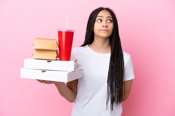 Poster - Teenager girl with braids holding pizzas and burgers over isolated pink background and looking up