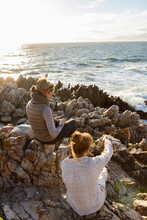 Woman And Teenage Girl Sitting On Rocky Shore, Looking Out To Sea.
