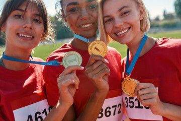 Portrait of excited diverse young female athletes showing their gold medals while standing together on race track