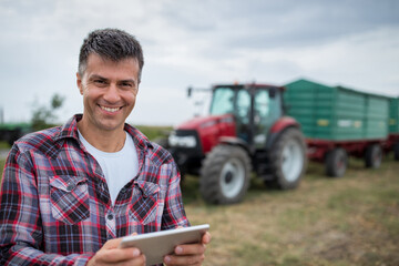 Wall Mural - Portrait of happy farmer holding tablet standing in front of tractor with trailer.