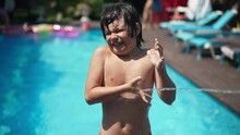 Medium Shot Portrait Of Confident Cute Caucasian Little Boy Standing At Swimming Pool With Hands On Hips As Water Splashing From Water Gun In Slow Motion. Playful Joyful Kid Having Fun On Vacations