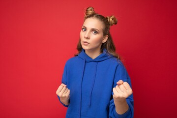 Photo of emotional angry young beautiful blonde woman with two horns with sincere emotions wearing hipster bright blue hoodie isolated over red background with free space and ready to fight