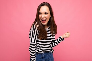 Portrait of young positive happy attractive brunette woman with sincere emotions wearing casual striped pullover isolated on pink background with free space and celebrating winning shouting yeah