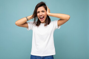 Portrait of emotional young attractive brunette woman with sincere emotions wearing casual white t-shirt for mockup isolated on blue background with empty space and covering ears with hands and