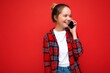 Attractive cute positive smiling young blonde woman standing isolated over red wall wearing casual red shirt and white t-shirt talking on mobile phone looking to the side