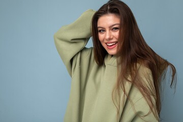 Wall Mural - Portrait of young beautiful smiling girl in stylish hipster green hoodie. Sexy carefree woman posing near blue wall. Positive model having fun