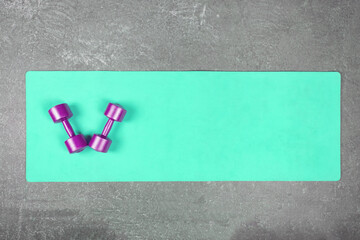 Flat lay of a turquoise yoga mat with violet dumbbells on the concrete floor. Pair of dumbbells on fitness mat spread out on cement floor