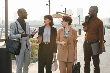 Horizontal medium long shot of four young multi-ethnic men and women finishing workday strolling along street together drinking coffee and chatting