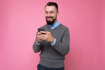 Photo shot of handsome positive good looking young man wearing casual stylish outfit poising isolated on background with empty space holding in hand and using mobile phone messaging sms looking at