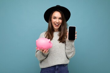 Wall Mural - Portrait photo of happy positive smiling satisfied sincere young attractive brunette woman wearing stylish grey sweater and black hat isolated on blue background with free space and holding pink pig