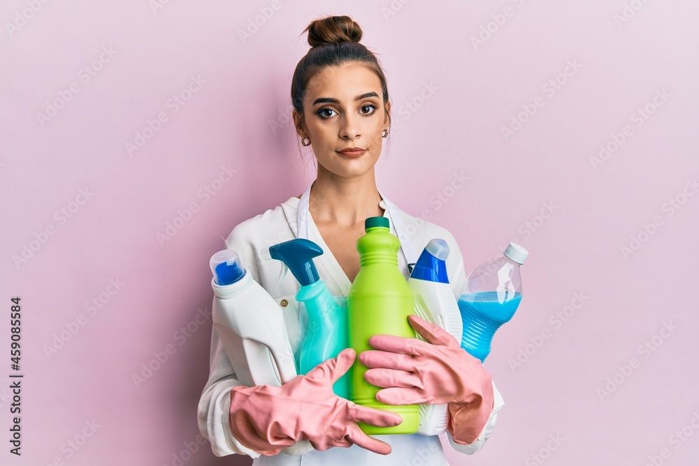 Beautiful brunette young woman wearing cleaner apron holding cleaning products relaxed with serious expression on face. simple and natural looking at the camera.
