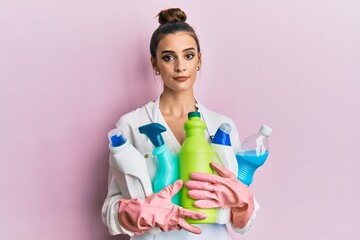 Wall Mural - Beautiful brunette young woman wearing cleaner apron holding cleaning products relaxed with serious expression on face. simple and natural looking at the camera.