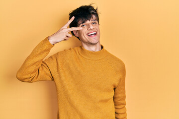 Handsome hipster young man wearing casual yellow sweater doing peace symbol with fingers over face, smiling cheerful showing victory